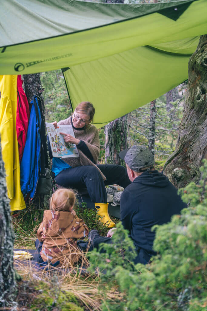 Nainen lukee lapsille kirjaa ulkona tarppikankaan alla.
